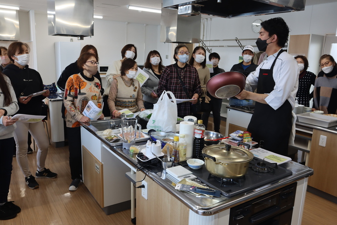 写真：調理室内で、調理器具を手に新メニューをつくる上でのポイント説明する講師と、その周りに集まって真剣に話しを聞く参加者