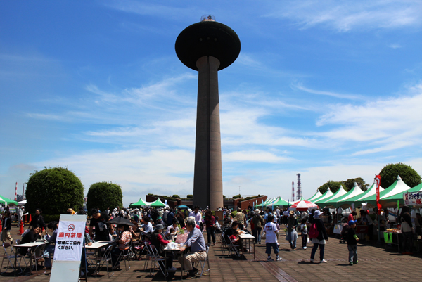 写真：晴天のもと、展望塔の下に集まる人々と港公園の様子