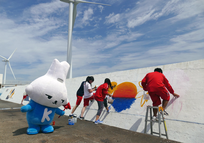 写真：白い壁面に、青い海に沈む夕日を描く1000人画廊制作中の高校生四人と応援するカミスココくん