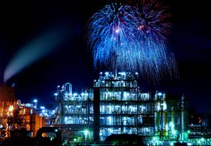写真：光り輝く夜の工場群の後ろに、大きな青い花火がふたつ。光の共演です。