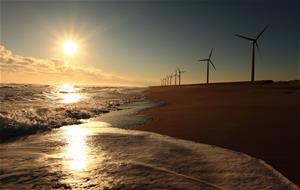 写真：左に波打ち際、右に風力発電が並ぶ海辺で、まばゆい光りとともに朝日が昇る「静刻」