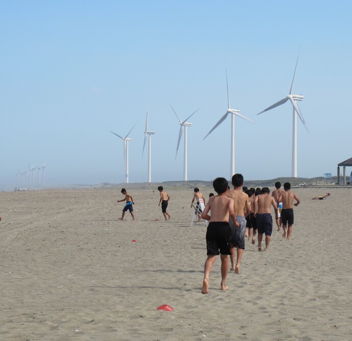 写真：遠くに並ぶ風力発電施設を背景に、砂浜でランニングする選手たち