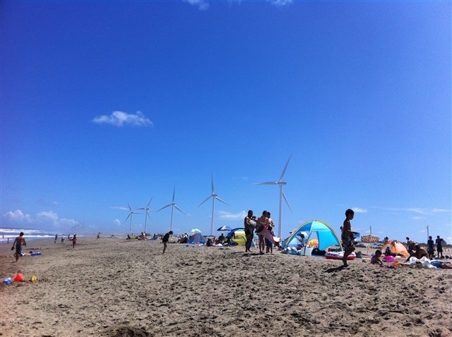 写真：青空のもと、海水浴客でにぎわう砂浜と風車