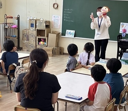 写真：学級訪問のようす。栄養士が人形を用いて給食についてのお話をしていて、それを聞き入る園児たち
