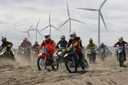 写真：風車を背にバイクに乗った参加者がビーチで競い合う様子