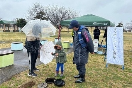 写真：神栖中央公園にて撮影。来場した親子にマンホールトイレについて説明する防災士