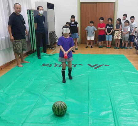 写真：目隠しをしてスイカ割りをしている女の子と、そのようすを見ている大人と子どもたち