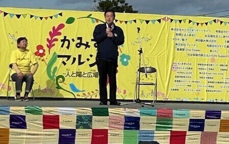 写真：2024年11月16日撮影。かみすマルシェの黄色い横断幕があるステージ上にて挨拶する市長