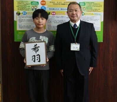 写真：やたべ土合小学校にて撮影。「希羽」という文字が書かれたパネルを持つ子どもと学校の先生
