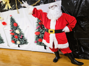 写真：サンタクロースが壁に装飾されたクリスマスツリーの前でポーズをとる様子